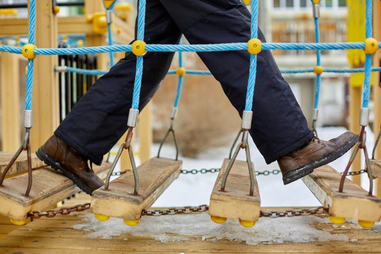 Close-up of child's legs walking on a wooden and rope bridge at a snowy playground.